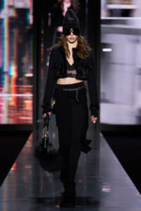 Model walks a fashion runway in an all-black, edgy look: cropped fur jacket, lace bralette, black pants with a chain belt, beanie, and large sunglasses, carrying a black fluffy handbag.