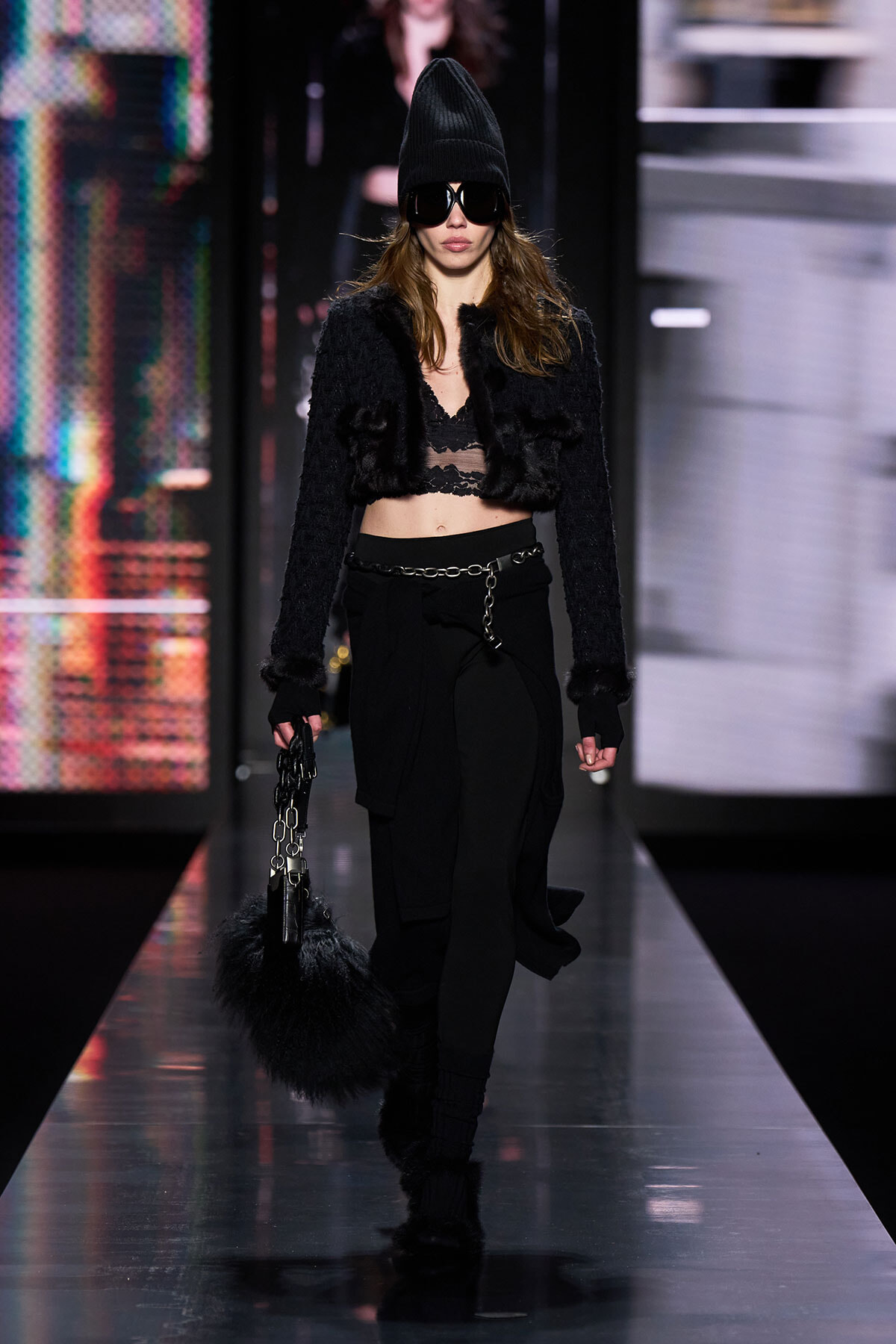 Model walks a fashion runway in an all-black, edgy look: cropped fur jacket, lace bralette, black pants with a chain belt, beanie, and large sunglasses, carrying a black fluffy handbag.