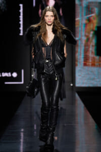Model walks a fashion runway in a black leather outfit with fur-trimmed shoulders, sheer top, and matching leather pants; carrying a black bag with chain straps.
