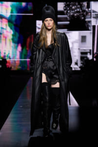 Model walking a fashion runway in a long black leather coat, black knit beanie, lace lingerie-style outfit, and over-the-knee boots on a colorful stage backdrop