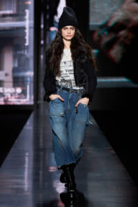 Model walks a fashion runway wearing a black beanie, cropped fur-trimmed jacket, graphic white tee, and oversized denim cargo jeans with black boots.