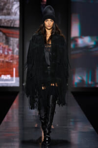 Model walking a fashion show in an all-black outfit: fringed suede coat, lace top, knit beanie, and tall buckled boots.