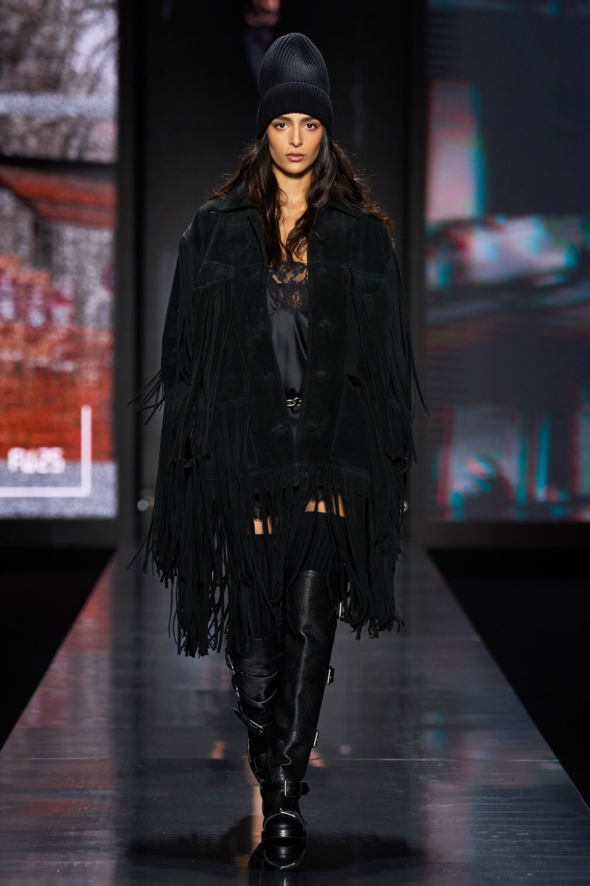 Model walking a fashion show in an all-black outfit: fringed suede coat, lace top, knit beanie, and tall buckled boots.