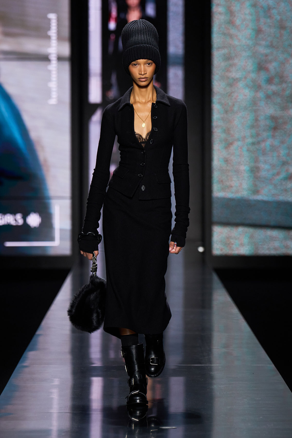 Model walking the runway in a black knit beanie, fitted black blazer, and long black skirt, carrying a furry black bag.