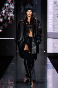 Model walking a runway in a black leather coat over a sheer lace top, black shorts, thigh-high socks, and chunky knee-high boots, wearing a black beanie
