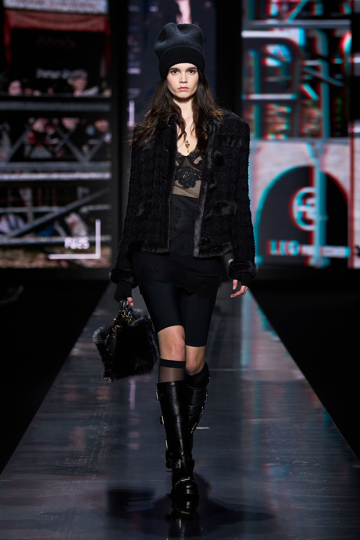 Female model walking a fashion runway in all-black ensemble: knit beanie, faux fur jacket, lace top, black shorts, tights, knee-high buckled boots, and a furry handbag.