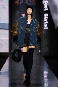 Model walking a fashion runway in a oversized denim jacket with leopard fur sleeves, black lace dress, and thigh-high socks.