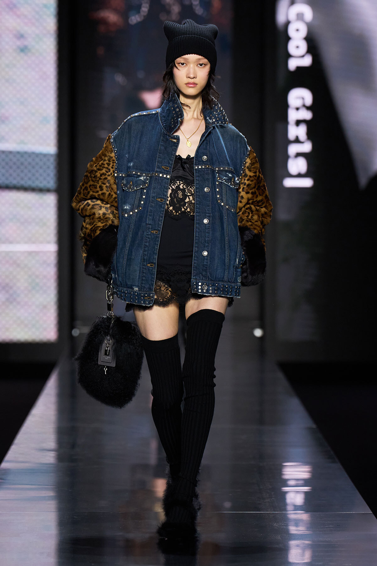 Model walking a fashion runway in a oversized denim jacket with leopard fur sleeves, black lace dress, and thigh-high socks.