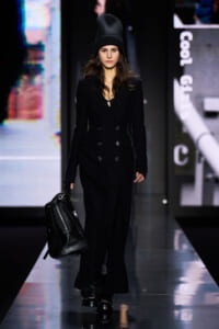 Model walks a fashion runway in an all-black ensemble: double-breasted coat, knit beanie, and a black crocodile-embossed bag in hand
