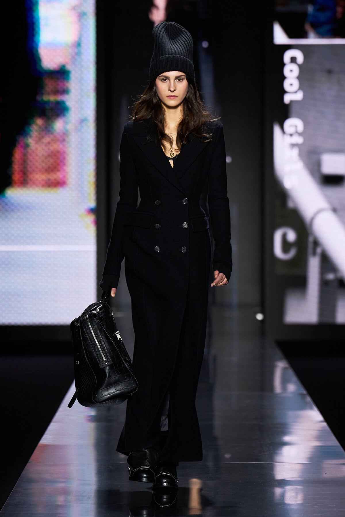 Model walks a fashion runway in an all-black ensemble: double-breasted coat, knit beanie, and a black crocodile-embossed bag in hand