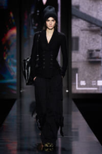 Female model walking a runway in an all-black outfit: double-breasted blazer, cargo pants, black beanie, and a large black bag over the shoulder.