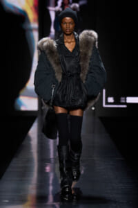 Model walking the runway in a black fur-trimmed coat, beanie, and tall buckled boots.