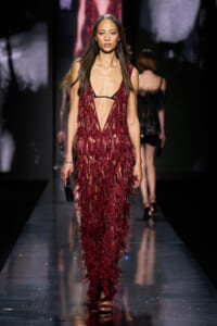 Runway model in a burgundy fringe gown with a deep V neckline, exposing a black bra, while carrying a small black clutch.