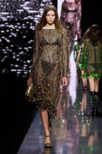Model walks the runway in a sheer gold-sequined gown with a gold clutch accessory.