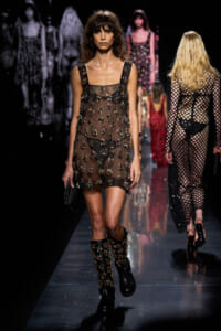 Model walking a fashion runway in a sheer black dress adorned with floral embellishments and matching boots, holding a small clutch.