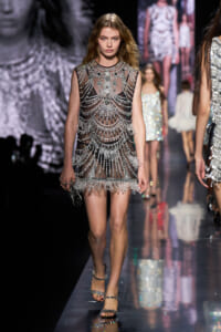 Model walking the runway in a sheer black dress covered with silver beaded chandeliers and lace trimming.