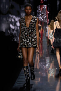 Model walks a jeweled black mini dress with floral sequins on a runway, carrying a small clutch and wearing embellished boots.