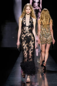 Model walks a fashion runway wearing a sheer black lace gown with a feathered hem and thin straps, holding a small clutch.