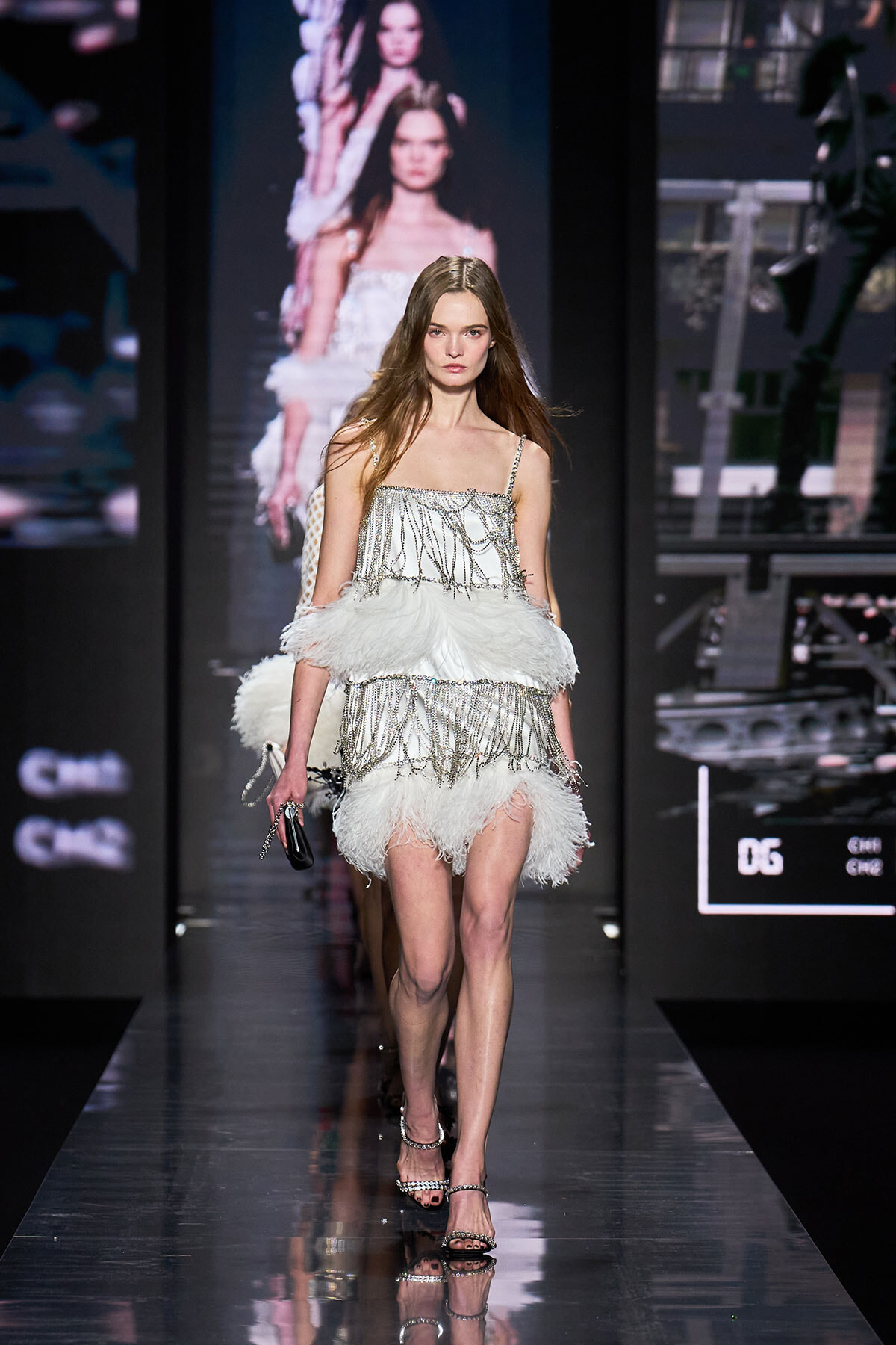 Model walks a glossy runway wearing a white feathered fringe dress with silver chain embellishments and strappy heels at a fashion show
