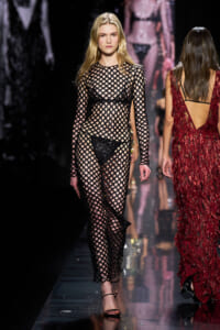 Model walking a fashion runway in a black fishnet dress over lingerie with a lattice pattern on the sheer fabric.