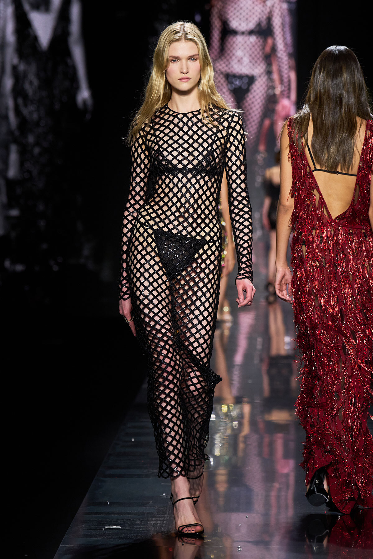 Model walking a fashion runway in a black fishnet dress over lingerie with a lattice pattern on the sheer fabric.