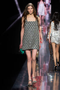 Model walking runway in a sleeveless, black-and-silver patterned mini dress with geometric floral motifs, paired with silver jeweled sandals and a small green clutch visible at her side.