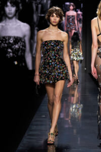 Model walking the runway in a strapless black mini dress covered with multicolored circular gems, paired with jeweled sandals.