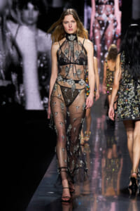 Model walking the runway in a sheer black mesh gown with silver floral appliqués and black lingerie visible beneath it.