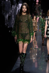 Model walks a fashion runway in a sheer green beaded mini dress with embellished boots.