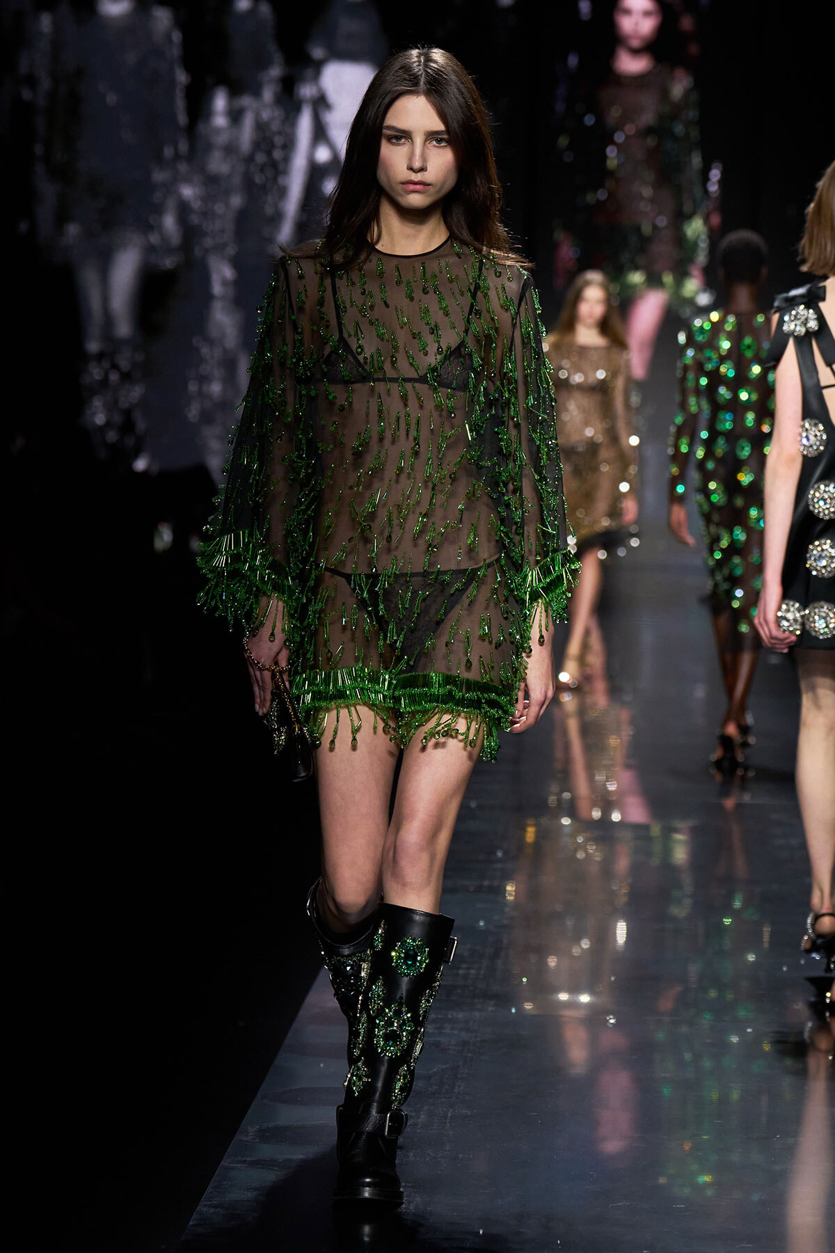 Model walks a fashion runway in a sheer green beaded mini dress with embellished boots.