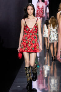 Model walks the runway in a red embellished mini dress with gold jewel appliques and thin straps, paired with jeweled black boots and a red clutch.