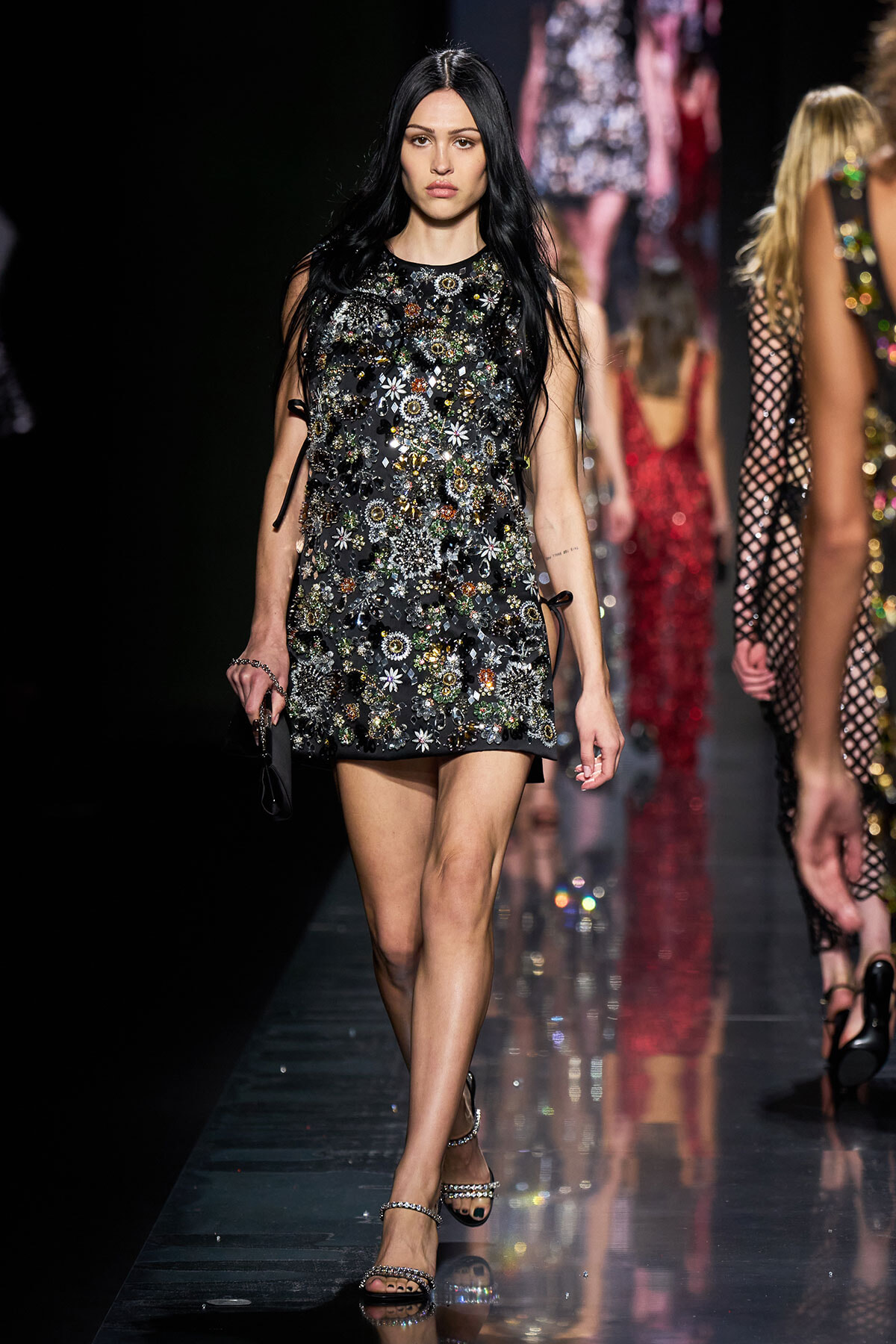 Runway model walking the catwalk in a black mini dress covered in colorful jeweled floral appliqués and strappy sandals.
