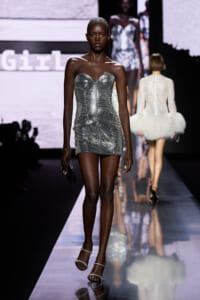 Model walks the runway in a silver, strapless, sequined mini dress with a small black clutch and sparkly sandals on a glossy catwalk.