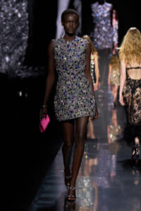 Model walks the runway in a sleeveless, gem-covered silver mini dress, paired with a pink clutch and jeweled sandals.