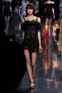 Model walks the runway in a black sequined mini dress with a dramatic feathered neckline.