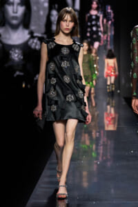 Model walking the runway in a black satin mini dress adorned with oversized silver jewel clusters and bow accents.