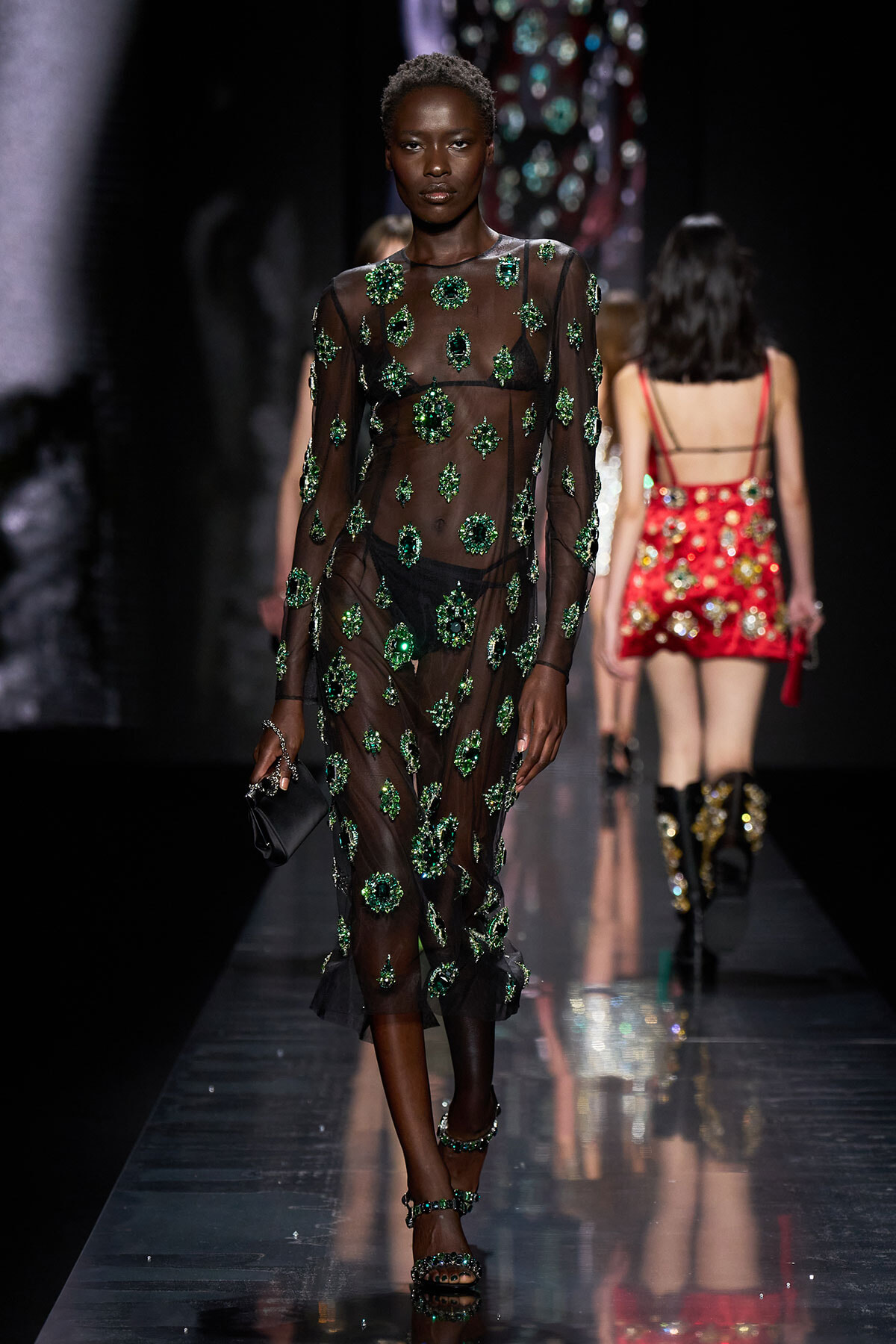 Model walks the runway in a sheer black dress covered in emerald-green crystals, clutch in hand.