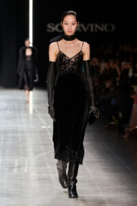 Model walking the runway in a black lace slip dress with sheer bodice, long black gloves, and knee-high boots.