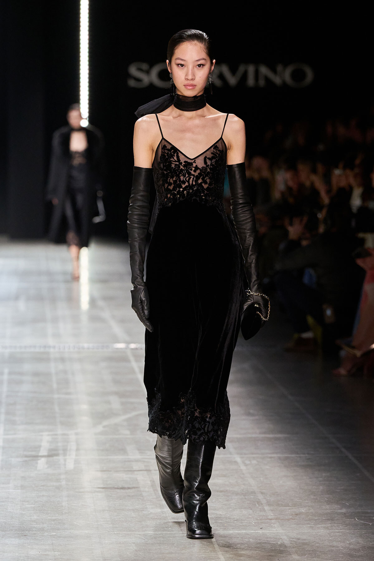 Model walking the runway in a black lace slip dress with sheer bodice, long black gloves, and knee-high boots.