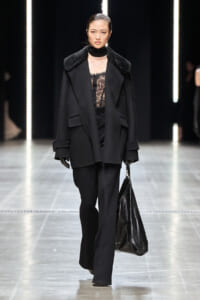 Model walking a runway in an all-black outfit: oversized coat with a fur collar, lace blouse, wide-leg pants, and a large black handbag.