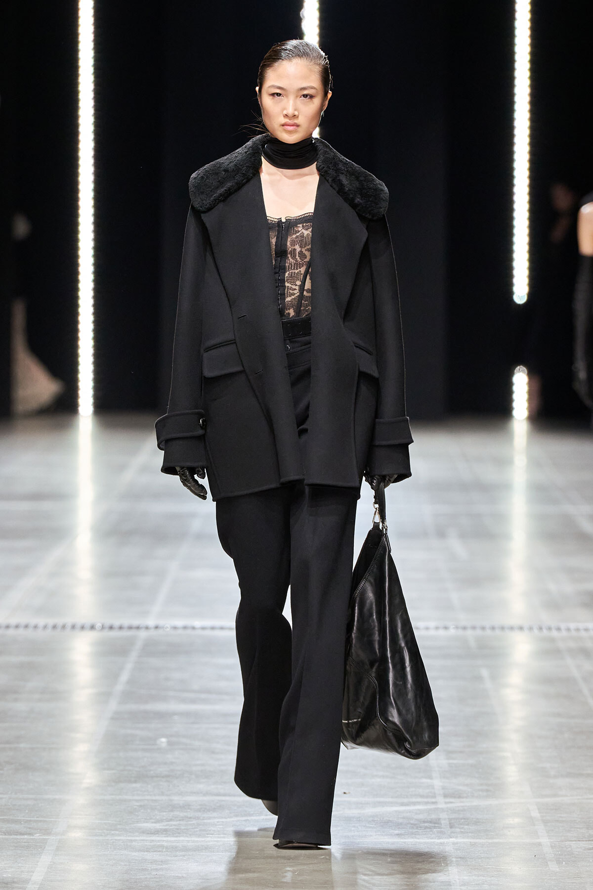 Model walking a runway in an all-black outfit: oversized coat with a fur collar, lace blouse, wide-leg pants, and a large black handbag.