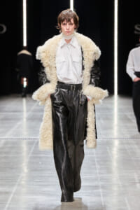 Model on a runway wearing a white button-down shirt, black leather pants, and an oversized cream shearling coat with a belt.