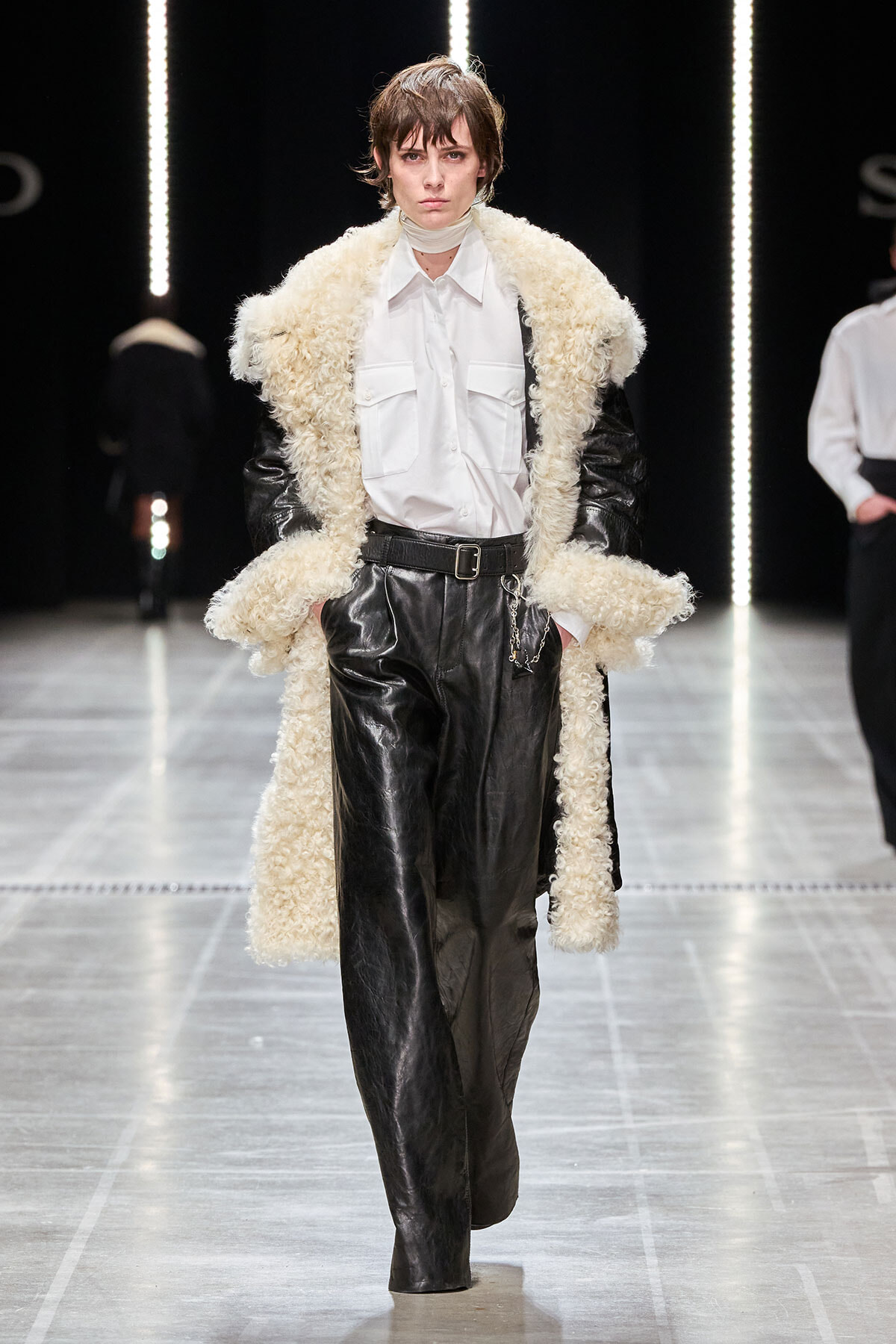 Model on a runway wearing a white button-down shirt, black leather pants, and an oversized cream shearling coat with a belt.