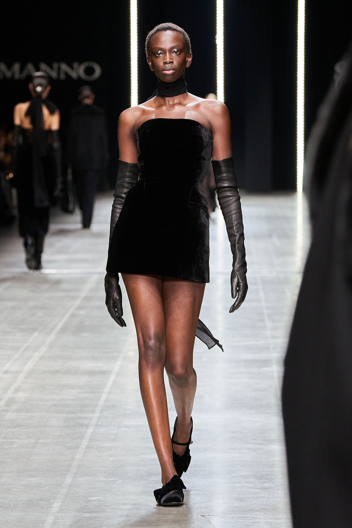 Model walks a black velvet strapless mini dress with long black gloves and choker on a runway.