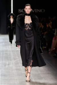 Model walks the runway in a long black coat with a plush fur collar, over a sheer black lace dress and black ankle-strap heels, carrying a glossy bag