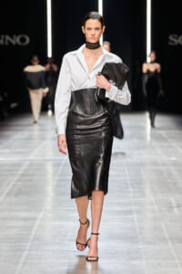 Model walking a fashion runway in a white blouse, black leather corset belt, and black leather midi skirt; carrying a black clutch and wearing strappy heels