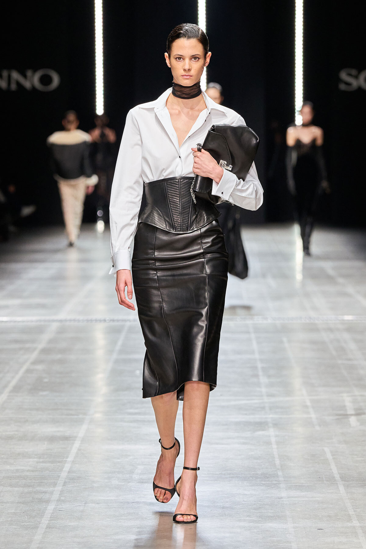 Model walking a fashion runway in a white blouse, black leather corset belt, and black leather midi skirt; carrying a black clutch and wearing strappy heels