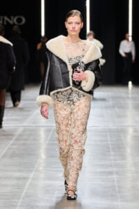 Model on a runway wearing a black shearling-edged leather jacket over a cream lace dress, carrying a black patterned clutch.
