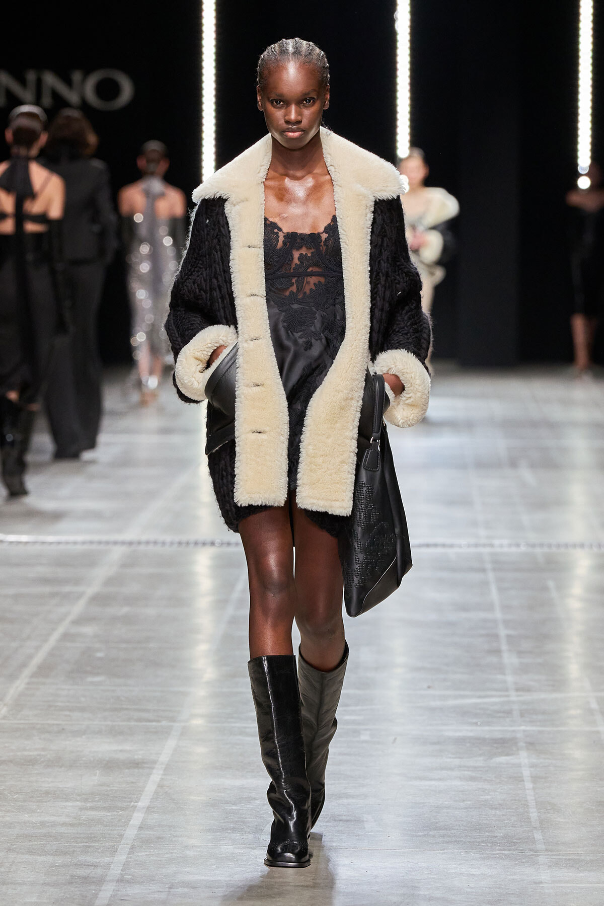 Model walking the runway in a black lace slip dress with a cream shearling coat and knee‑high black boots, carrying a black bag.