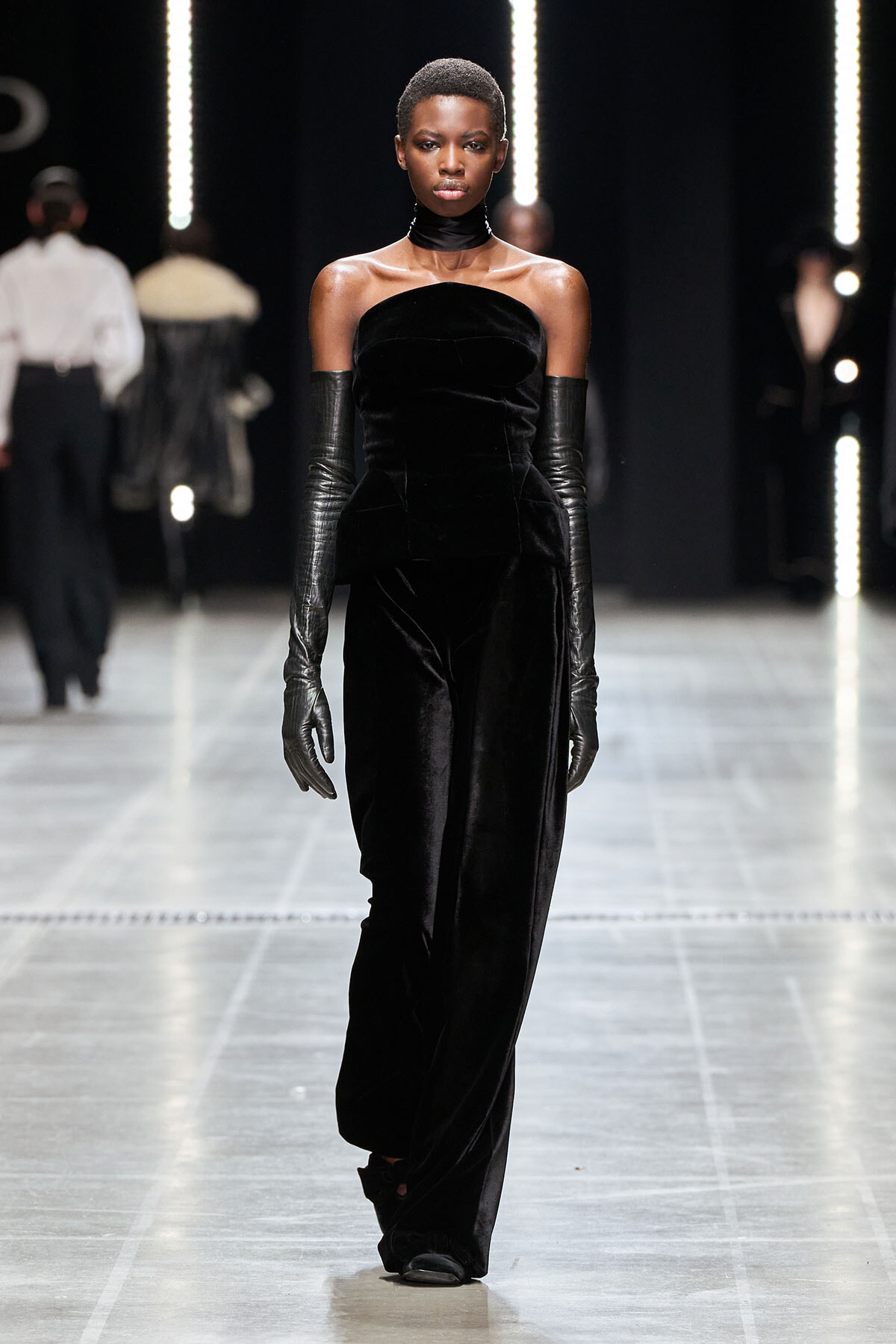 Runway model wearing a black velvet strapless jumpsuit with long matching gloves and a black choker.